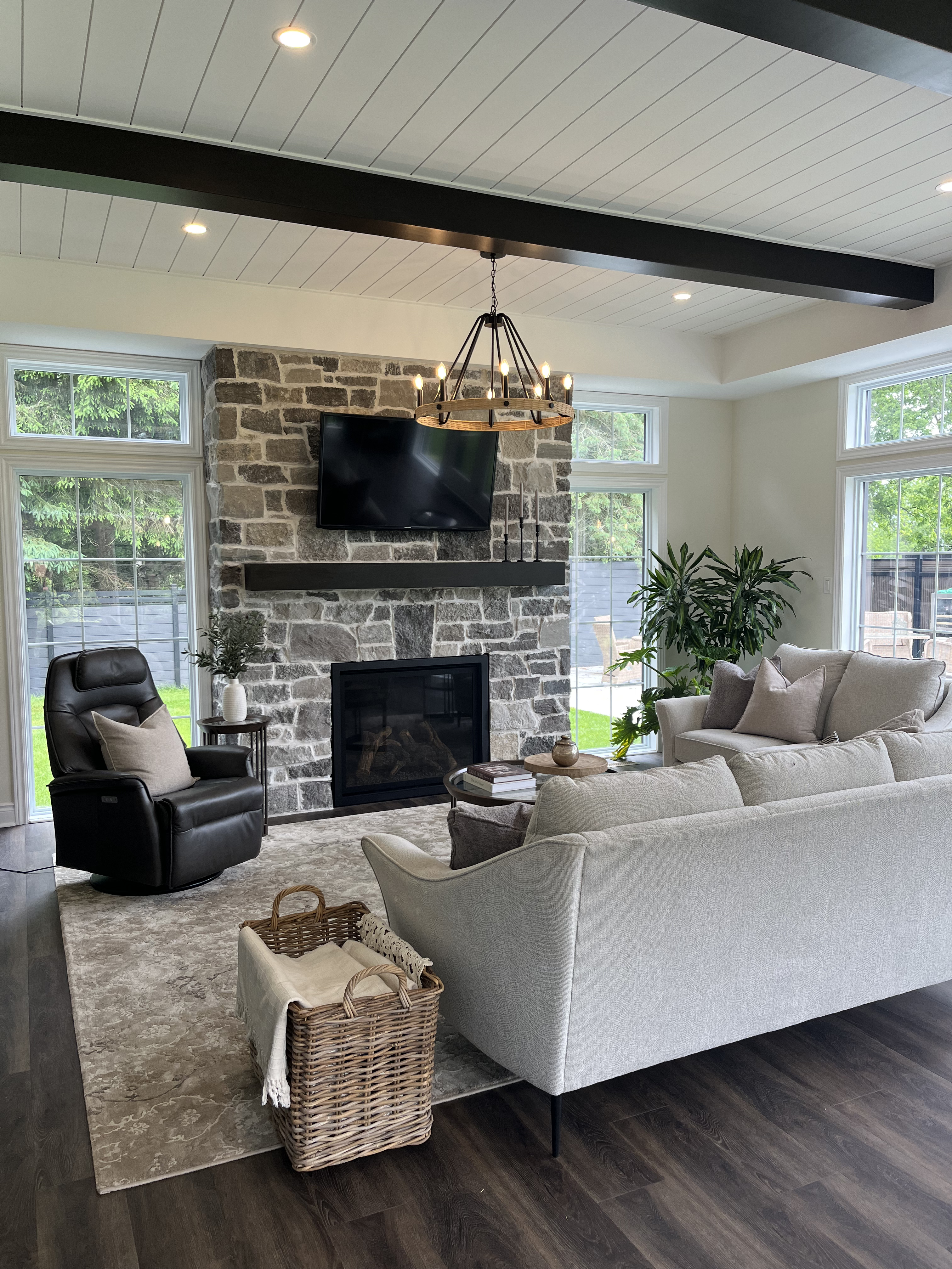 Modern home addition living room with stone fireplace, white shiplap ceiling, black ceiling beams, and cozy neutral furnishings, featuring a large window view.